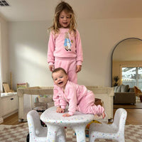 Two children in pink outfits playing on a small table in a home setting, with a hand holding a cup of coffee in the foreground.